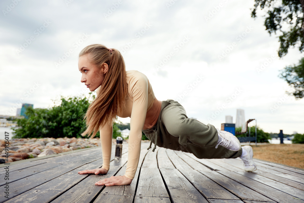 A beautiful woman, blonde, fitness instructor, in light clothes, trains on the beach, against the backdrop of the city. Shows exercises from yoga, pilates, deep work, step aerobics.
