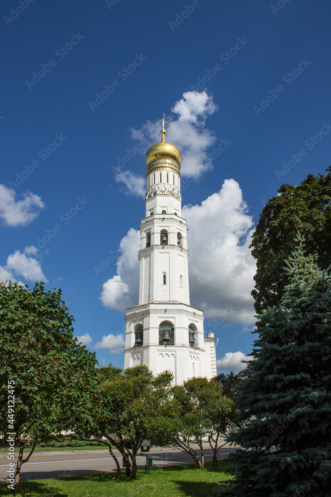 MOSCOW, RUSSIA-AUGUST, 4, 2021: the high white-stone bell tower of Ivan the Great with a golden dome against the background of blue sky and green foliage of a tree on a sunny summer day