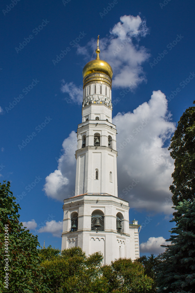 The high Kremlin white bell tower with a golden dome Ivan the Great in a natural frame made of tree branches with green leaves on a sunny summer day and blue sky in Moscow Russia