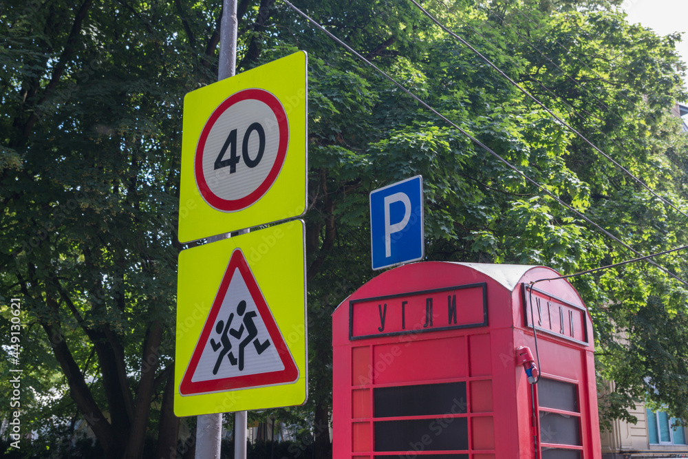 Road signs Caution, children and Speed limit 40 on pole with wires, next to them red booth with inscription in russian COALS, behind it blue parking sign against background of dense foliage of trees. Road signs Caution, children and Speed limit 40 on pole with wires, next to them red booth with inscription in russian COALS, behind it blue parking sign against background of dense foliage of trees.