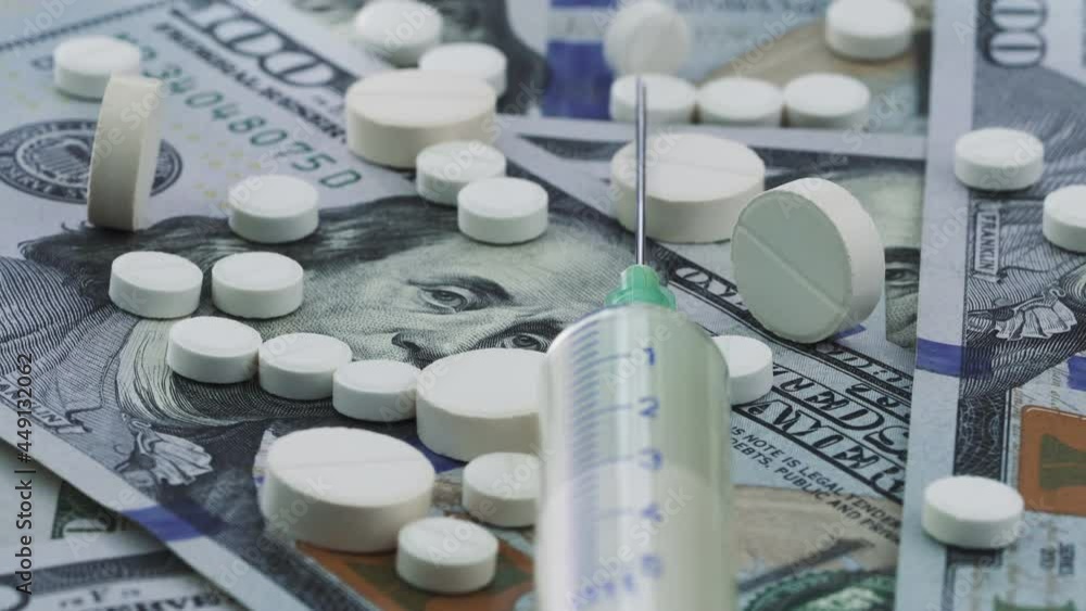 Close-up view of white medical pills and syringe rotating on the US dollars. The rising cost of health care. Pharmaceutical industry, medicine, addiction concepts. Close-up view of white medical pills and syringe rotating on the US dollars. The rising cost of health care. Pharmaceutical industry, medicine, addiction concepts.
