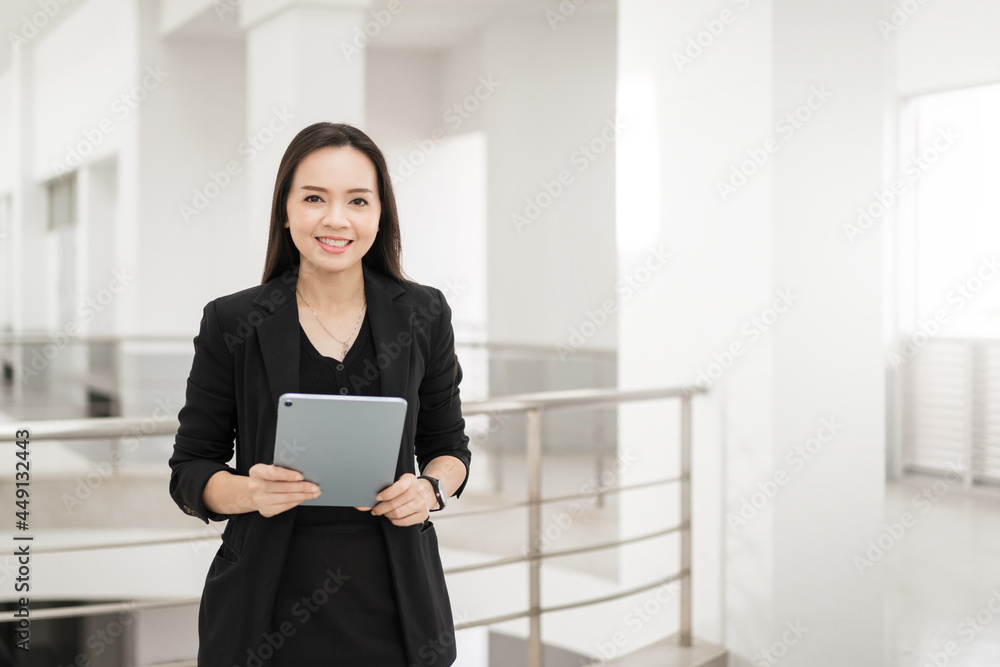 Portrait stock photo of a confident cheerful Asian businesswoman professional in black business suit using a digital tablet standing in the office building representing business oriented concept Portrait stock photo of a confident cheerful Asian businesswoman professional in black business suit using a digital tablet standing in the office building representing business oriented concept