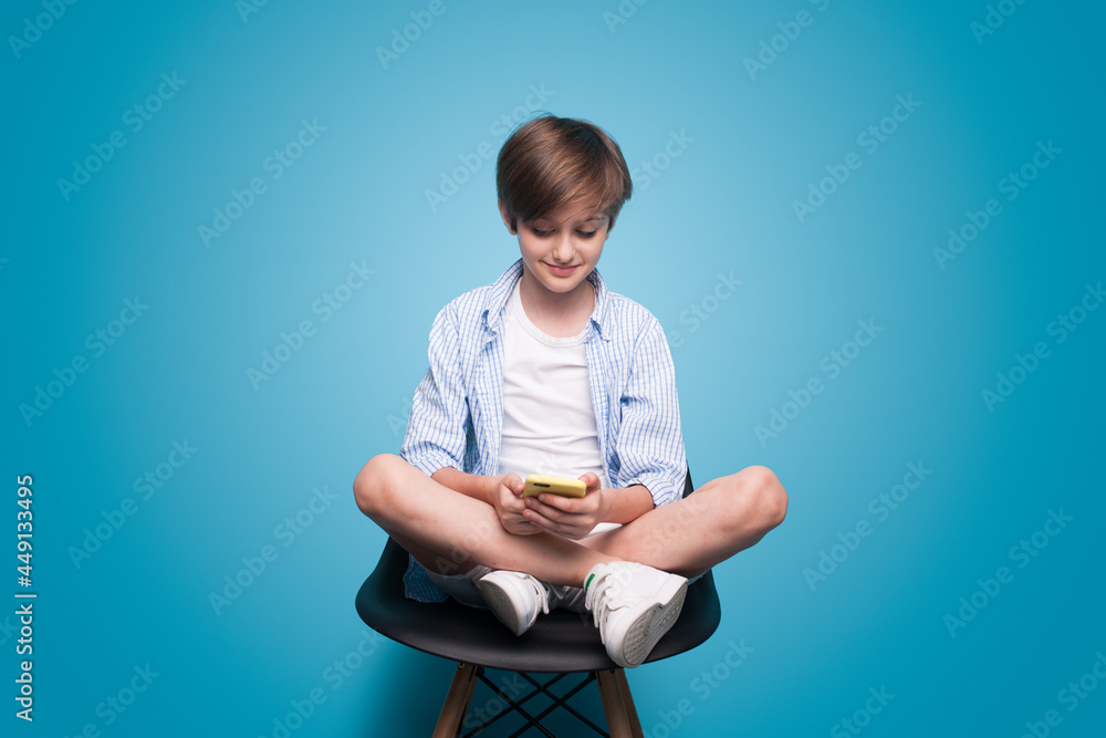 Caucasian schoolboy is sitting in armchair and using a phone on a blue studio wall Caucasian schoolboy is sitting in armchair and using a phone on a blue studio wall