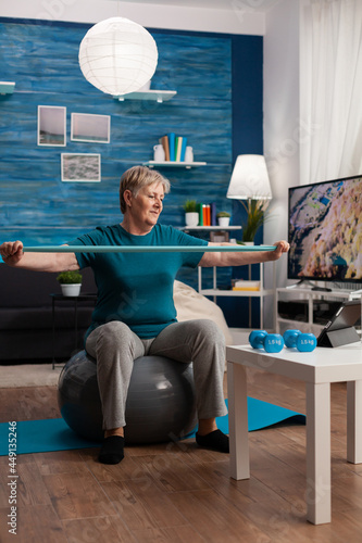 Cheerful retired pensioner working arm muscles using elastic band practicing aerobic exercise. Pensioner sitting on swiss ball in living room working at body healthcare resistance watching gym video photo