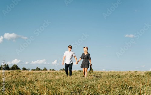 Happy beautiful couple of lovers holding hands walking in field against background blue sky, smiling people in love enjoying outdoor relaxation and time together. Romantic lifestyle, relationships © Sergio