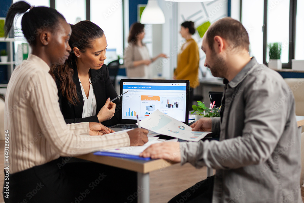 Busy multicultural, diverse employees analysing annual financial statistics sitting at desk in front of laptop holding documents searching business solutions. Team of businesspeople working in company Busy multicultural, diverse employees analysing annual financial statistics sitting at desk in front of laptop holding documents searching business solutions. Team of businesspeople working in company