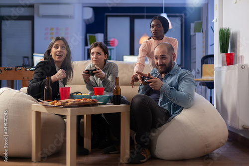 Multi ethnic group of coworkers play game on console while holding joystick. Cheerful diverse friends enjoy fun competition on tv computer while having snacks and beer of table