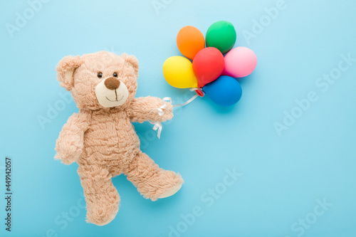 Smiling brown teddy bear holding heap of colorful balloons on light blue table background. Pastel color. Closeup. Congratulation concept. Top down view. photo