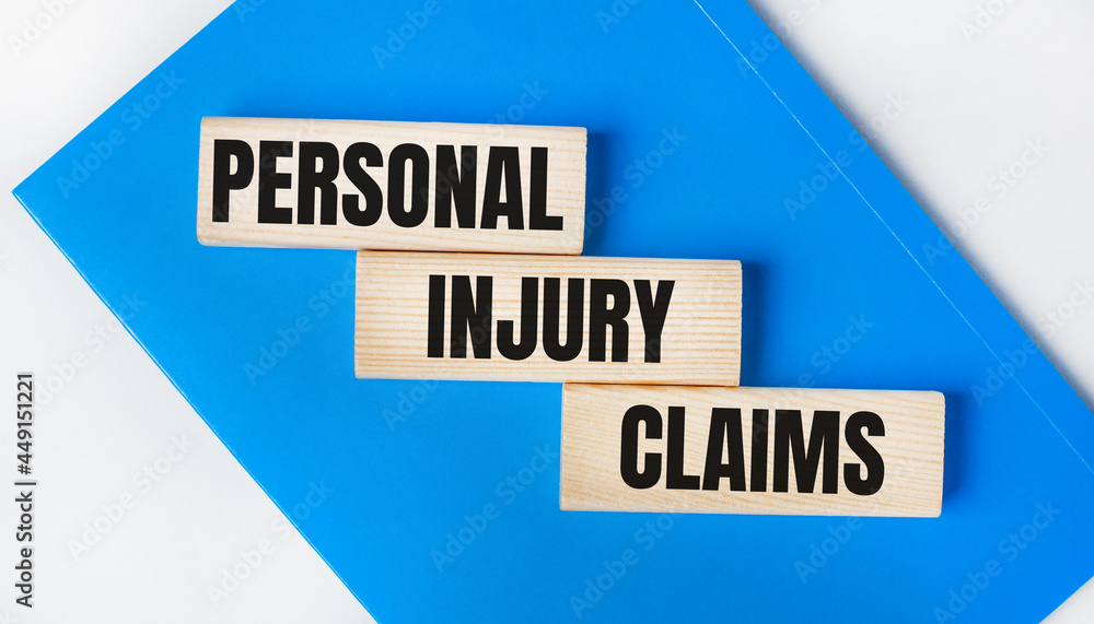 There is a blue notebook on a light gray background. Above are three wooden blocks with the words PERSONAL INJURY CLAIMS. There is a blue notebook on a light gray background. Above are three wooden blocks with the words PERSONAL INJURY CLAIMS.