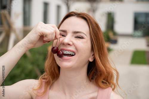 Beautiful young red-haired woman with braces on her teeth eats sweet cherries in the summer outdoors