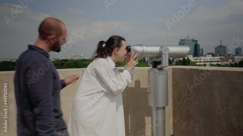 Caucasian tourist looking at panoramic view of metropolis using telescope on sky tower top. Young woman at observation point watching city and nature in metropolitan landscape