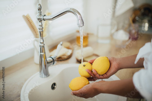 Young woman washing fresh lemons in a sink at the kitchen. Making lemonade. Apartment living.