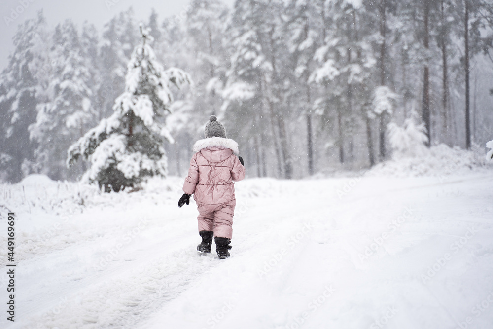 Cute little girl in pink jumpsuit is playing in the snow during a snowfall