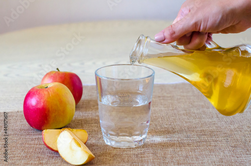 A woman's hand pours apple cider vinegar in a glass bottle on a light background into a glass of water. Malic acid is beneficial for health and is used in cooking