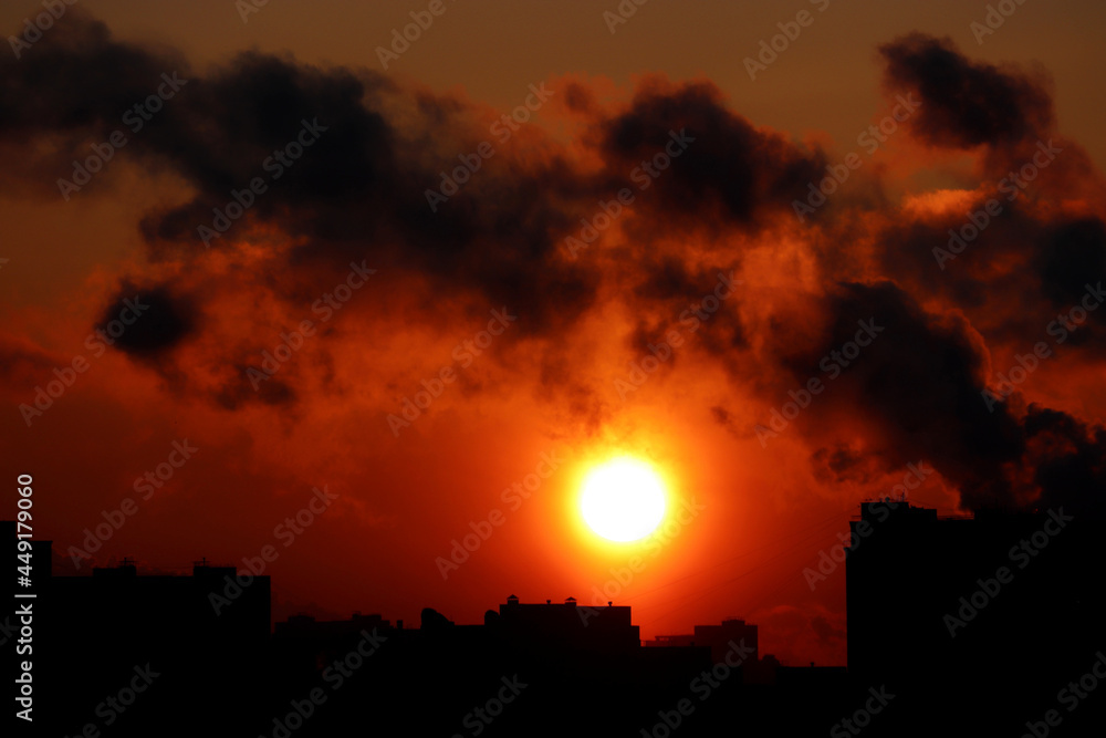 Fire in the city, orange sun shining through dark clouds of smoke above the buildings silhouettes
