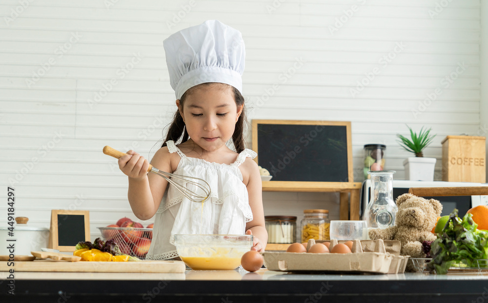 Cute daughter wearing chef's hat, having fun, cracking eggs, baking, mixing batter, cakes and chickpeas before microwave in home kitchen. Cute daughter wearing chef's hat, having fun, cracking eggs, baking, mixing batter, cakes and chickpeas before microwave in home kitchen.