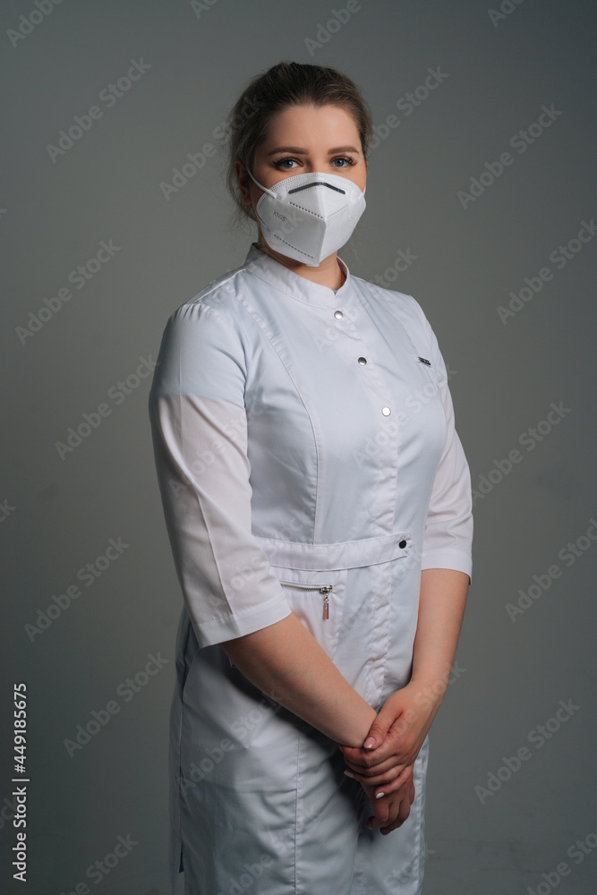 Vertical portrait of young female doctor wearing medical coat and protective face mask standing on dark isolated background. Studio shot of nurse posing looking at camera. Vertical portrait of young female doctor wearing medical coat and protective face mask standing on dark isolated background. Studio shot of nurse posing looking at camera.