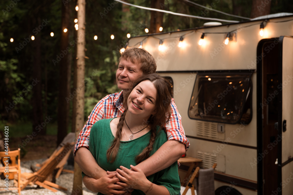 Couple camping in trailer park. Happy blond man hugging pretty woman close to mobile home parked in forest. Summer vacation in tiny house on wheels