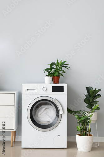 Modern washing machine with laundry and houseplants near light wall