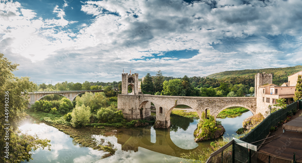 Besalu, Girona, Catalonia, Spain. Famous Landmark Old Medieval Romanesque Besalu Bridge Over The Fluvia River In Cloudy Summer Day