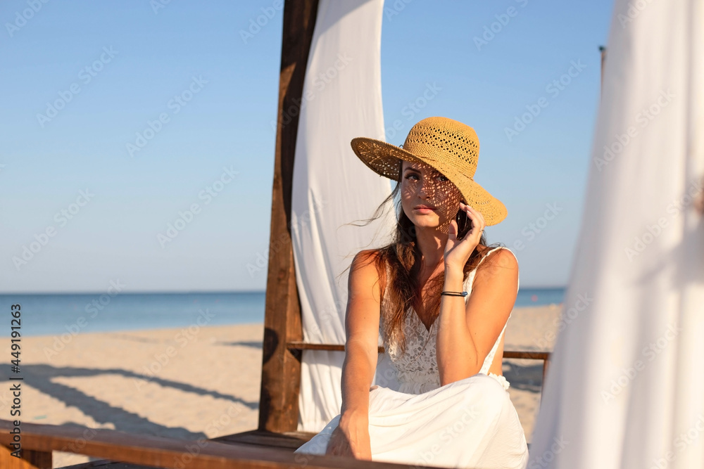A young girl in a white sundress and a straw hat, sitting resting, relaxing, meditating in a gazebo, a bungalow on the beach.
