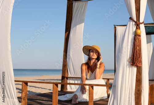 Foto A young girl in a white sundress and a straw hat, sitting resting, relaxing, meditating in a gazebo, a bungalow on the beach