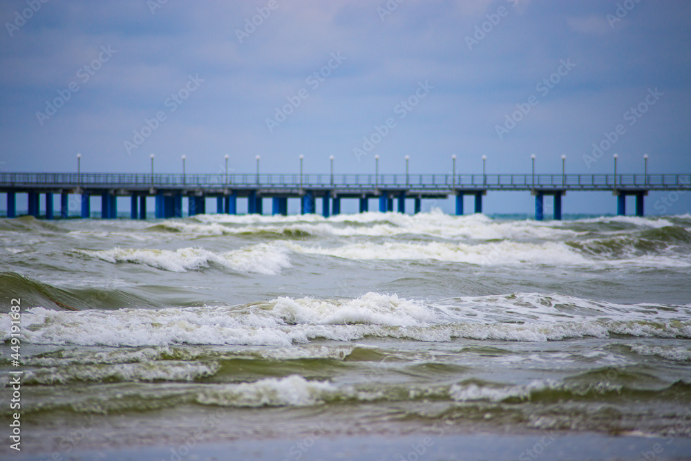 Seashore with gray-green waves and foam. Gloomy weather. The pier is visible in the background. Copy space.