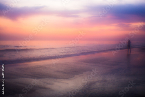 Mystical seascape at sunset. Fabulous haze and pink-violet colors. A silhouette of a girl is walking along the sandy shore along the water. Copy space.