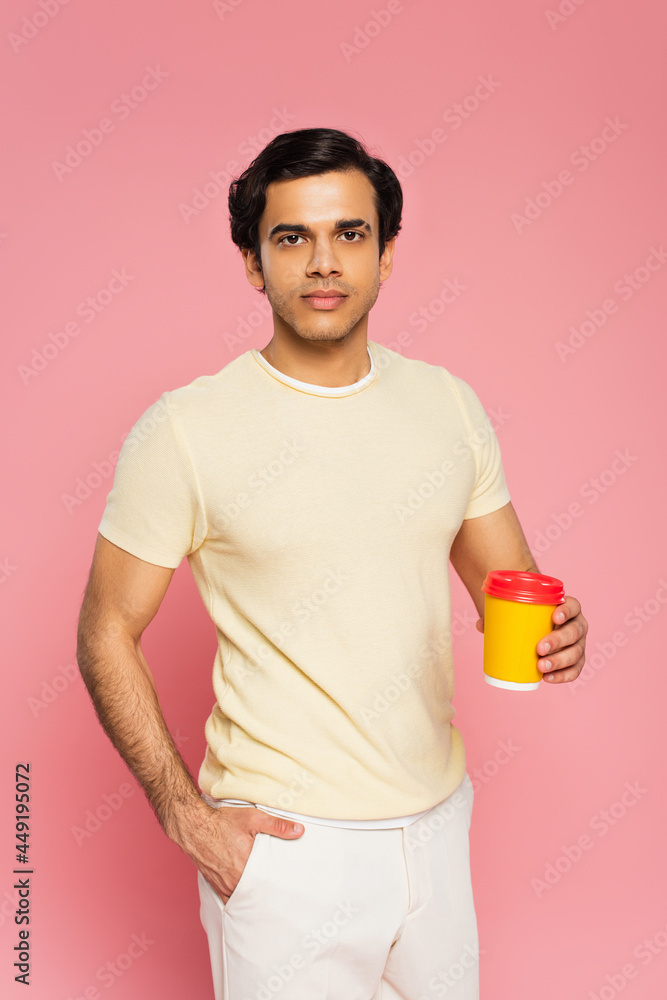 young man holding paper cup with coffee to go while standing with hand in pocket isolated on pink young man holding paper cup with coffee to go while standing with hand in pocket isolated on pink