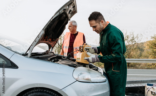 Two road assistant workers in towing service trying to start car engine with jump starter and energy station with air compressor. Roadside assistance concept.