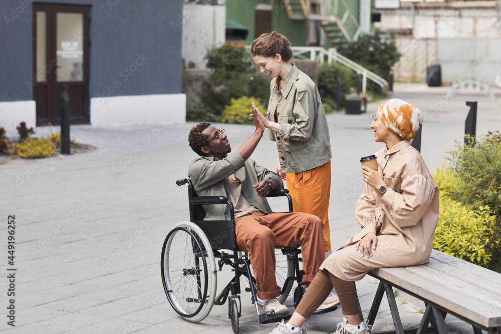 Obraz premium Happy young Caucasian woman giving five to disabled black man in wheelchair while welcoming him outdoors