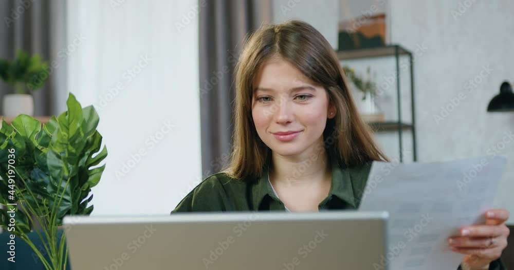 View over the laptop on good-looking satisfied smiling skilful young female freelancer which sitting at her workplace and checking information with report and laptop View over the laptop on good-looking satisfied smiling skilful young female freelancer which sitting at her workplace and checking information with report and laptop