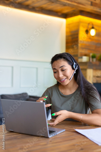 Portrait of a smiling asian woman with headset in front of laptop monitor.