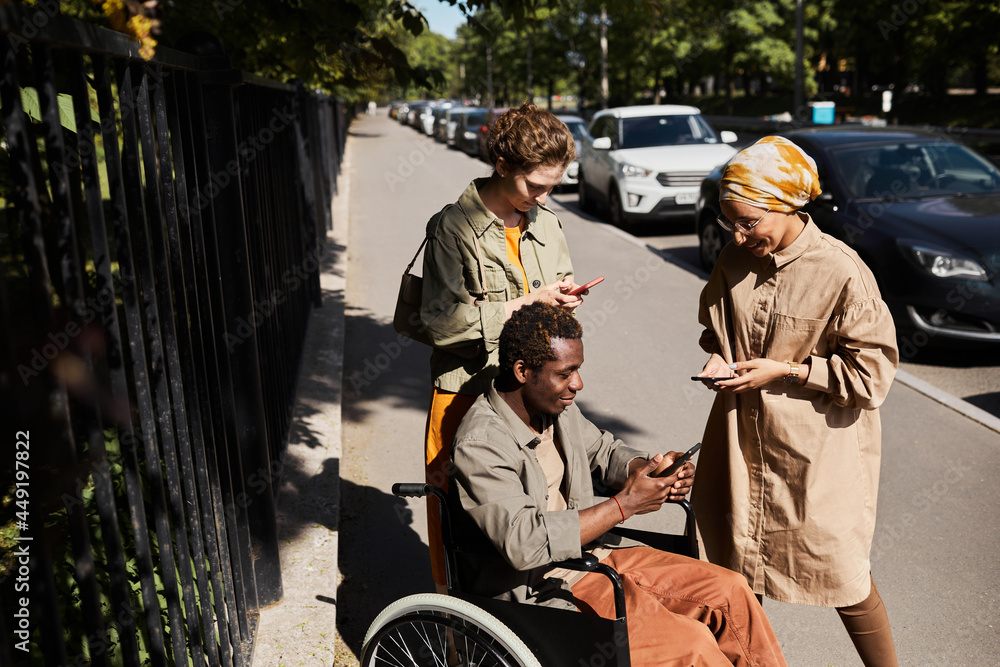 Group of young multi-ethnic friends with disabled man in wheelchair using gadgets on street Group of young multi-ethnic friends with disabled man in wheelchair using gadgets on street