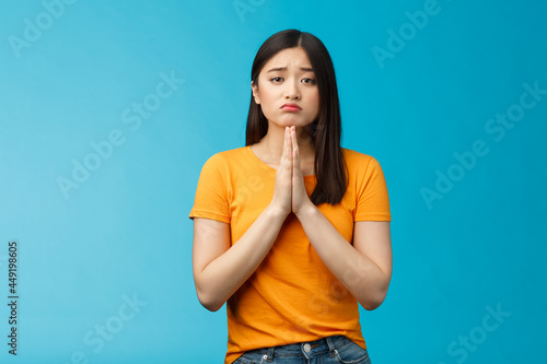 Upset cute silly asian girl praying, plead for help, pouting frowning need, make pitty face, hold hands pray begging for favor, apologizing feelings guilty sad, stand blue background photo
