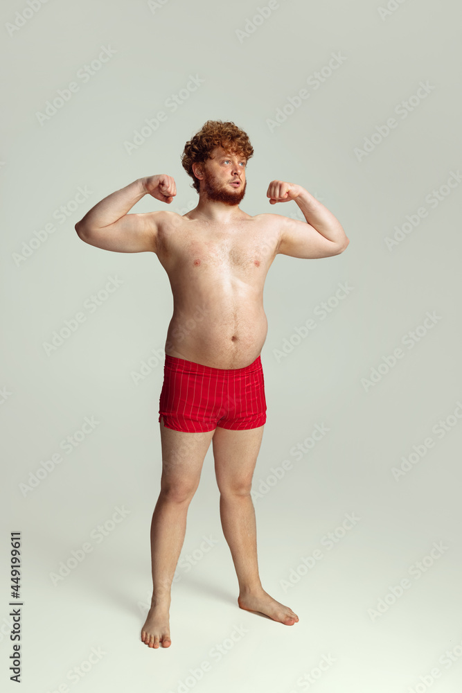 Cute red-headed man in red swimming shorts posing isolated on gray studio background. Concept of sport, humor and body positive.