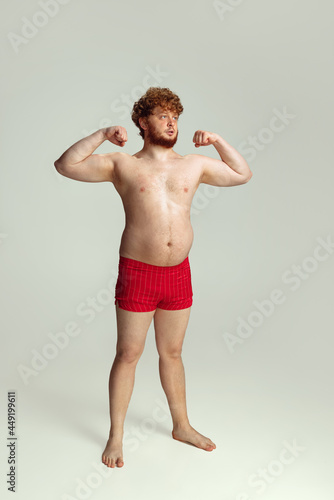 Cute red-headed man in red swimming shorts posing isolated on gray studio background. Concept of sport, humor and body positive. © master1305