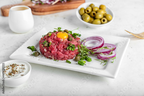 Plate with tasty beef tartare, olives and sauce on light background