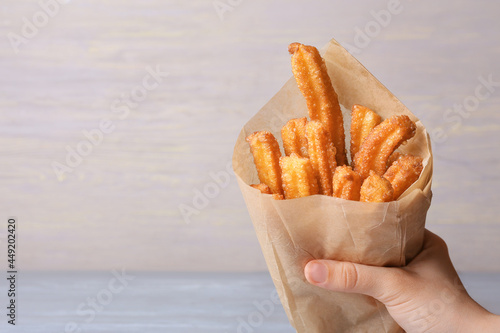 Female hand with tasty churros in parchment on light wooden background, closeup photo