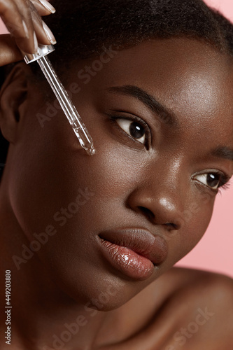 Portrait close up of beautiful black girl dropping serum collagen moisturizer on face. Serious young woman. Concept of face skin care. Isolated on pink background. Studio shoot
