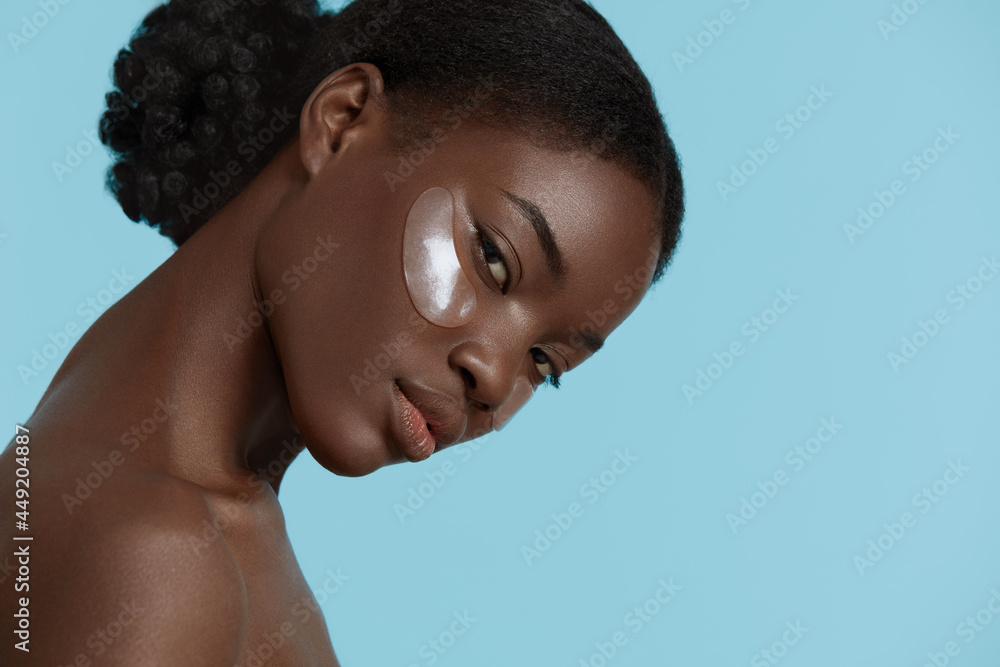 Close up portrait of beautiful african girl with under eye patch on face. Serious young woman looking at camera. Concept of face skin care. Isolated on blue background. Studio shoot