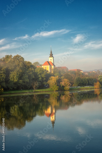 Scenic view of Zupnija Maribor Sv. Jozef church in misty morning landscape with colorful autumn trees, lush foliage, calm water of Drava river and reflection, blue sky with clouds, Maribor, Slovenia