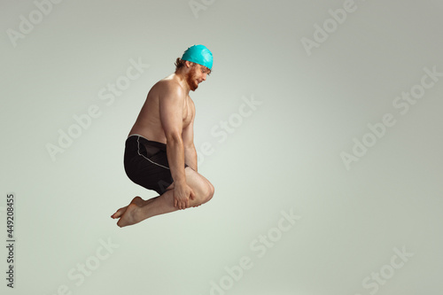 Funny dive. Cute red-headed man in red swimming shorts posing isolated on gray studio background. Concept of sport, humor and body positive.