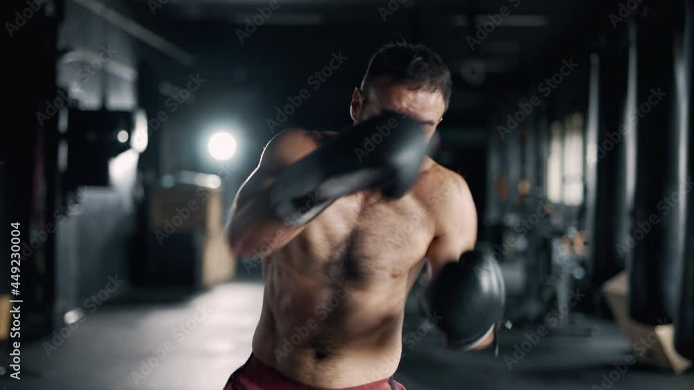 A muscular sportsman is boxing in the dark gym. Self confident boxer is looking at the camera and training indoors. Maximum focus and strength. Concept of lifestyle health and sports. Close up. A muscular sportsman is boxing in the dark gym. Self confident boxer is looking at the camera and training indoors. Maximum focus and strength. Concept of lifestyle health and sports. Close up.
