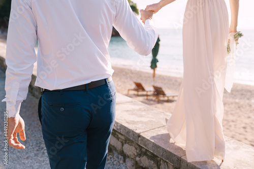 Groom holds the hand of bride with a bouquet, walking along the seashore