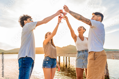 Group of multiracial happy friends toasting at the beach with red wine at sunset clinking glasses in front of sunlight in backlit. Young people having fun in outdoor party drinking alcohol together