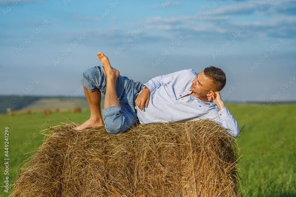 A young European man lies on a bale of hay in a loose, comfortable position. Portrait of a farmer in a shirt and jeans. Summer holiday on the farm. A young European man lies on a bale of hay in a loose, comfortable position. Portrait of a farmer in a shirt and jeans. Summer holiday on the farm.