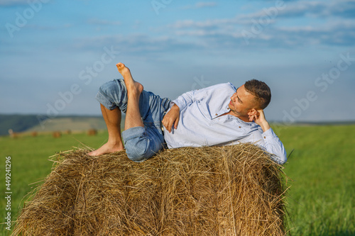 A young European man lies on a bale of hay in a loose, comfortable position. Portrait of a farmer in a shirt and jeans. Summer holiday on the farm.