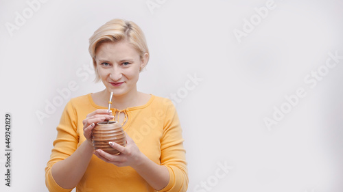 Beautiful girl holding a brown container shape like a coconut with a straw. About to drink with the straw. The girl is dressed in a yellow top. Isolated over white background. Tropical vacations.