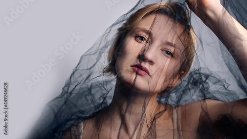 Fashion Model with a black net veil. Isolated woman under black cloth Net. Portrait of a blonde girl with a serious facial expression trying to escape. High-quality studio shot.